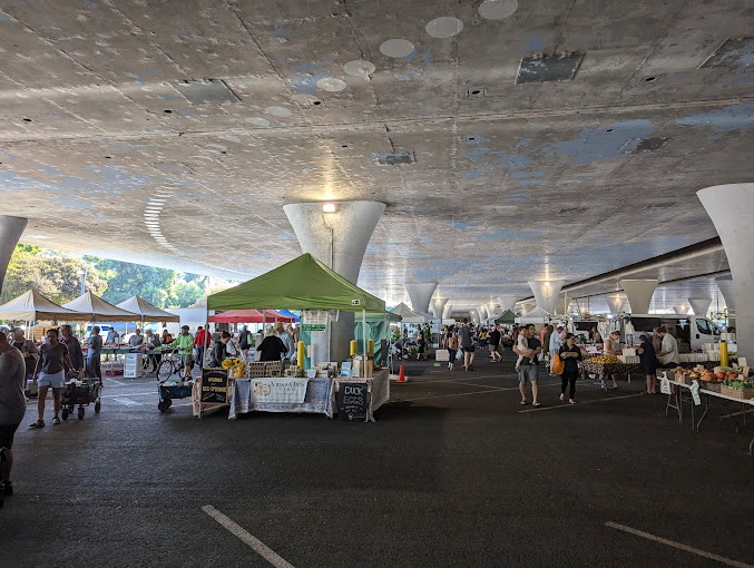 Two rows of farmers market canopy tents with customers walking between them and the light rail train climbing the hill in the background in front of a cloudy blue sky.